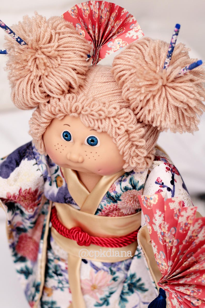 Op de afbeelding: Pop met blauwe ogen en sproeten, gekleed in een bloemenkimono met een rode touwriem. De pop heeft garen haar gestyled in twee grote knotjes en houdt een rode papieren waaier vast. Het haar van de pop is versierd met decoratieve stokjes.