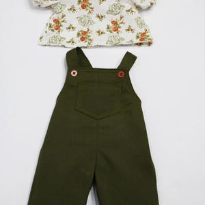 May include: A two-piece doll outfit featuring a floral print top and olive green overalls. The top has a white background with a floral pattern in shades of orange and green. The overalls have a front pocket and wooden buttons.