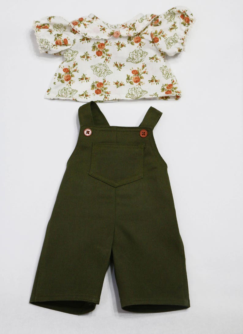 May include: A two-piece doll outfit featuring a floral print top and olive green overalls. The top has a white background with a floral pattern in shades of orange and green. The overalls have a front pocket and wooden buttons.