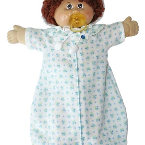 May include: A vintage doll with brown curly hair, freckles, and a dummy. The doll wears a white sleep sack with a blue floral pattern and a matching collar. The doll's arms are outstretched.