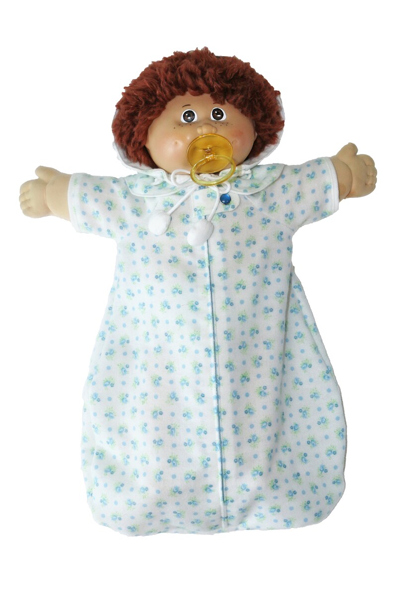 May include: A vintage doll with brown curly hair, freckles, and a dummy. The doll wears a white sleep sack with a blue floral pattern and a matching collar. The doll's arms are outstretched.