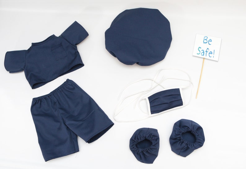 K&ouml;nnte beinhalten: Marineblaues Puppen-OP-Kittel-Set, einschlie&szlig;lich Oberteil, Shorts, M&uuml;tze, Gesichtsmaske und Schuh&uuml;berzieher. Ein kleines Schild mit "Be Safe!" ist ebenfalls enthalten. Die Artikel sind auf einer wei&szlig;en Oberfl&auml;che angeordnet.