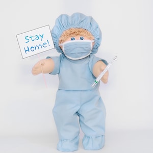K&ouml;nnte beinhalten: Eine Puppe in hellblauer medizinischer Kleidung, einer passenden M&uuml;tze und einer Gesichtsmaske. Die Puppe h&auml;lt ein Schild mit der Aufschrift "Stay Home!" und ein Thermometer. Die Puppe hat blondes Haar und blaue Augen.