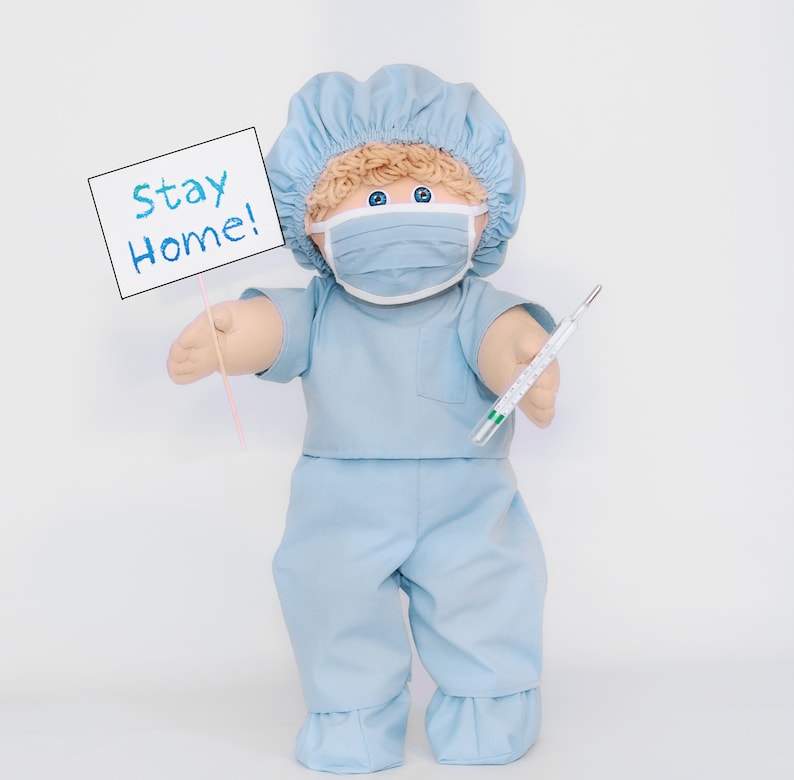 K&ouml;nnte beinhalten: Eine Puppe in hellblauer medizinischer Kleidung, einer passenden M&uuml;tze und einer Gesichtsmaske. Die Puppe h&auml;lt ein Schild mit der Aufschrift "Stay Home!" und ein Thermometer. Die Puppe hat blondes Haar und blaue Augen.