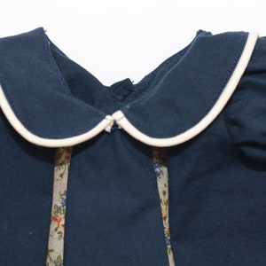 May include: A navy blue dress with a white-trimmed Peter Pan collar and ruffled sleeves. The dress features floral patterned ribbon accents down the front. The dress is made of a soft fabric and is suitable for children.