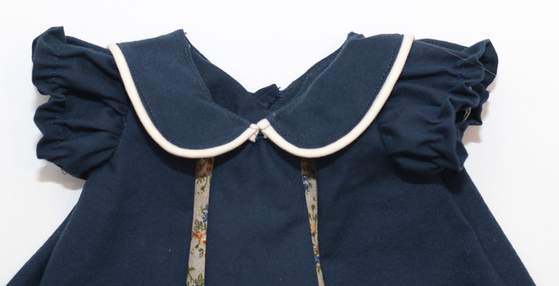 May include: A navy blue dress with a white-trimmed Peter Pan collar and ruffled sleeves. The dress features floral patterned ribbon accents down the front. The dress is made of a soft fabric and is suitable for children.