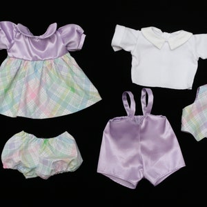 May include: A collection of doll clothes in pastel colors. The set includes a lavender dress with a plaid skirt, bloomers, a white shirt, suspender shorts, a vest, and a bow tie. The items are arranged on a black background.