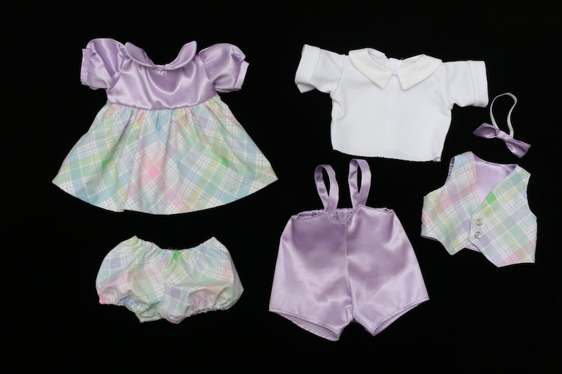 May include: A collection of doll clothes in pastel colors. The set includes a lavender dress with a plaid skirt, bloomers, a white shirt, suspender shorts, a vest, and a bow tie. The items are arranged on a black background.