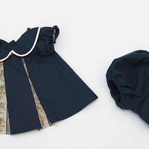 May include: Navy blue baby dress and bloomers set. The dress features a white-trimmed Peter Pan collar, short sleeves, and floral patterned panels. The bloomers are a matching navy blue color.