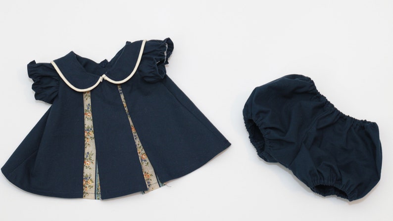 May include: Navy blue baby dress and bloomers set. The dress features a white-trimmed Peter Pan collar, short sleeves, and floral patterned panels. The bloomers are a matching navy blue color.