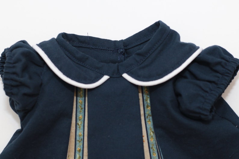 May include: Navy blue dress with a white-trimmed Peter Pan collar and short, puffed sleeves. The dress features vertical panels with a floral design in shades of blue, green, and yellow.