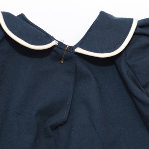 May include: A navy blue garment with a white-trimmed sailor collar and ruffled sleeves. A gold safety pin secures the front. The fabric appears to be a soft cotton, suitable for children's clothing.