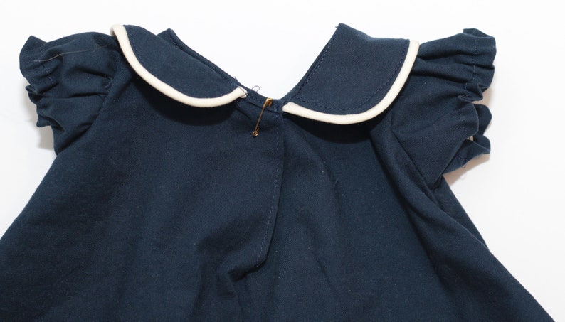 May include: A navy blue garment with a white-trimmed sailor collar and ruffled sleeves. A gold safety pin secures the front. The fabric appears to be a soft cotton, suitable for children's clothing.