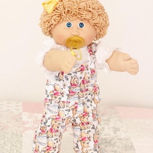 Könnte beinhalten: Eine Puppe mit lockigem, beigem Wollhaar, blauen Augen und einer gelben Haarschleife. Die Puppe trägt ein weißes Kurzarmshirt unter einer weißen Latzhose mit einem wiederkehrenden Muster von Winnie Puuh. Ein gelber Schnuller steckt im Mund.