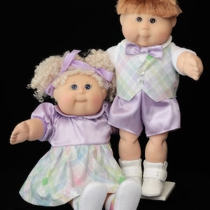 May include: Two Cabbage Patch Kids dolls. One doll is seated, wearing a lavender dress with a plaid skirt and white shoes. The other doll is standing, wearing a plaid vest, lavender shorts, and white shoes. Both dolls have light skin and brown eyes.