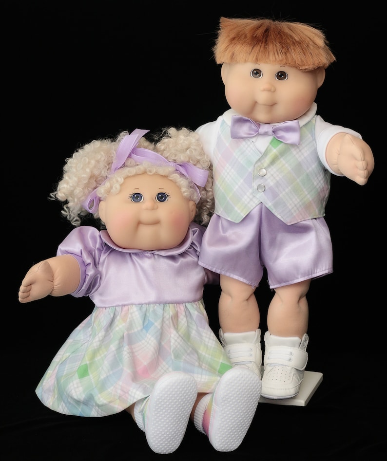 May include: Two Cabbage Patch Kids dolls. One doll is seated, wearing a lavender dress with a plaid skirt and white shoes. The other doll is standing, wearing a plaid vest, lavender shorts, and white shoes. Both dolls have light skin and brown eyes.
