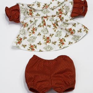 May include: A two-piece doll outfit featuring a white top with a floral pattern and a Peter Pan collar. The sleeves are a rust-colored fabric with a polka dot design. Matching rust-colored bloomers with a polka dot pattern complete the set.