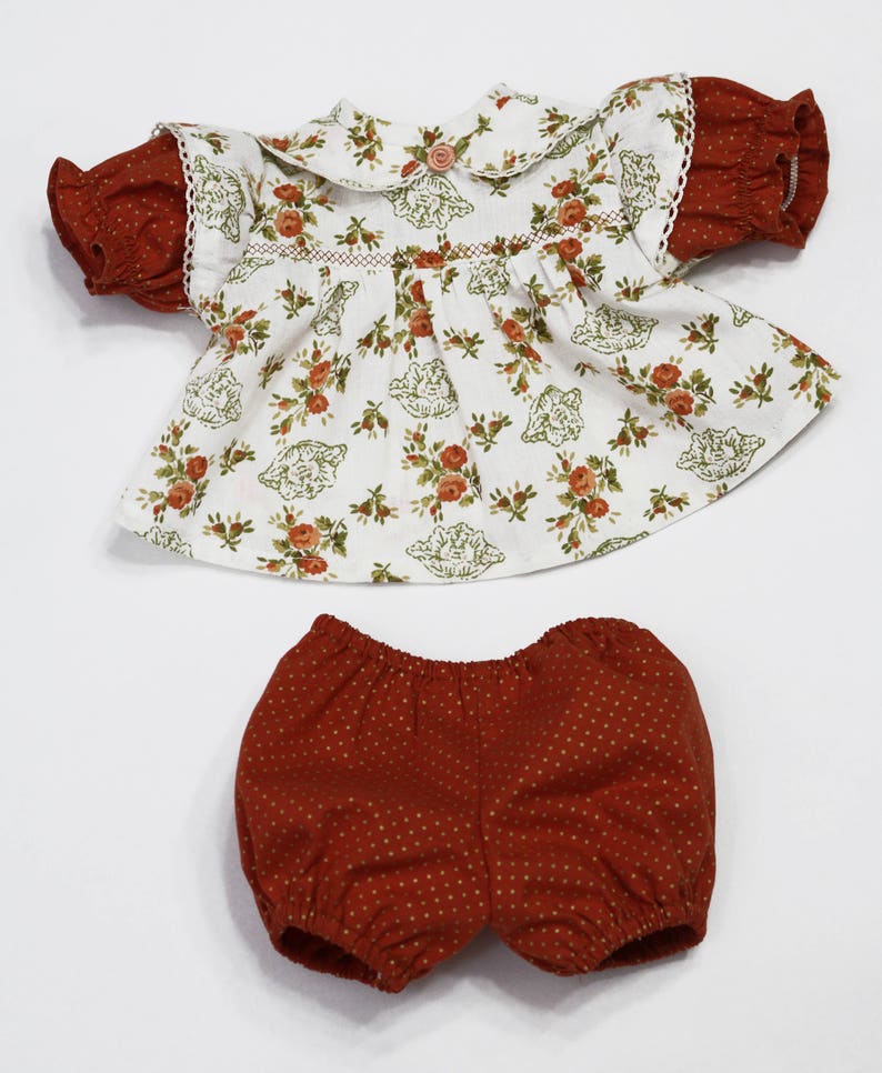 May include: A two-piece doll outfit featuring a white top with a floral pattern and a Peter Pan collar. The sleeves are a rust-colored fabric with a polka dot design. Matching rust-colored bloomers with a polka dot pattern complete the set.