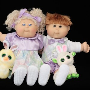 May include: Two Cabbage Patch Kids dolls, a girl and a boy, sit side-by-side. The girl doll has blonde curly hair and wears a lavender dress. The boy doll has brown hair and a plaid vest. Each doll holds a plush animal.