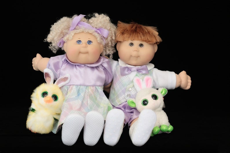May include: Two Cabbage Patch Kids dolls, a girl and a boy, sit side-by-side. The girl doll has blonde curly hair and wears a lavender dress. The boy doll has brown hair and a plaid vest. Each doll holds a plush animal.