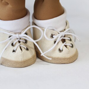 May include: Close-up of a pair of off-white canvas sneakers with white laces and metal eyelets. The shoes have a tan rubber sole and are worn with white socks and tan pants. The sneakers are likely for a doll or small child.