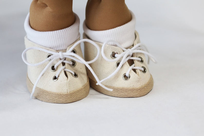 May include: Close-up of a pair of off-white canvas sneakers with white laces and metal eyelets. The shoes have a tan rubber sole and are worn with white socks and tan pants. The sneakers are likely for a doll or small child.