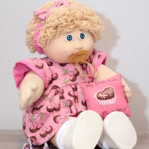May include: A vintage doll with blonde curly hair, blue eyes, and a pacifier. The doll wears a pink dress with a cherry pattern and holds a pink pillow with the text "My first love... Chocolate!"