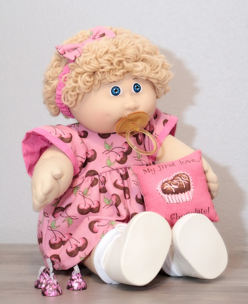 May include: A vintage doll with blonde curly hair, blue eyes, and a pacifier. The doll wears a pink dress with a cherry pattern and holds a pink pillow with the text "My first love... Chocolate!"