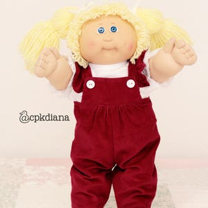 May include: A doll with light yellow yarn hair styled in pigtails, blue eyes, and rosy cheeks. The doll wears a white short-sleeved shirt with lace trim and dark red corduroy overalls featuring two white buttons. The text @cpkdiana is visible.