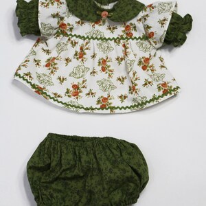 May include: A doll's outfit featuring a white top with a floral pattern in orange and green, a green collar, and matching green ruffled sleeves. The outfit includes green bloomers with a floral design. The outfit is designed for a doll.