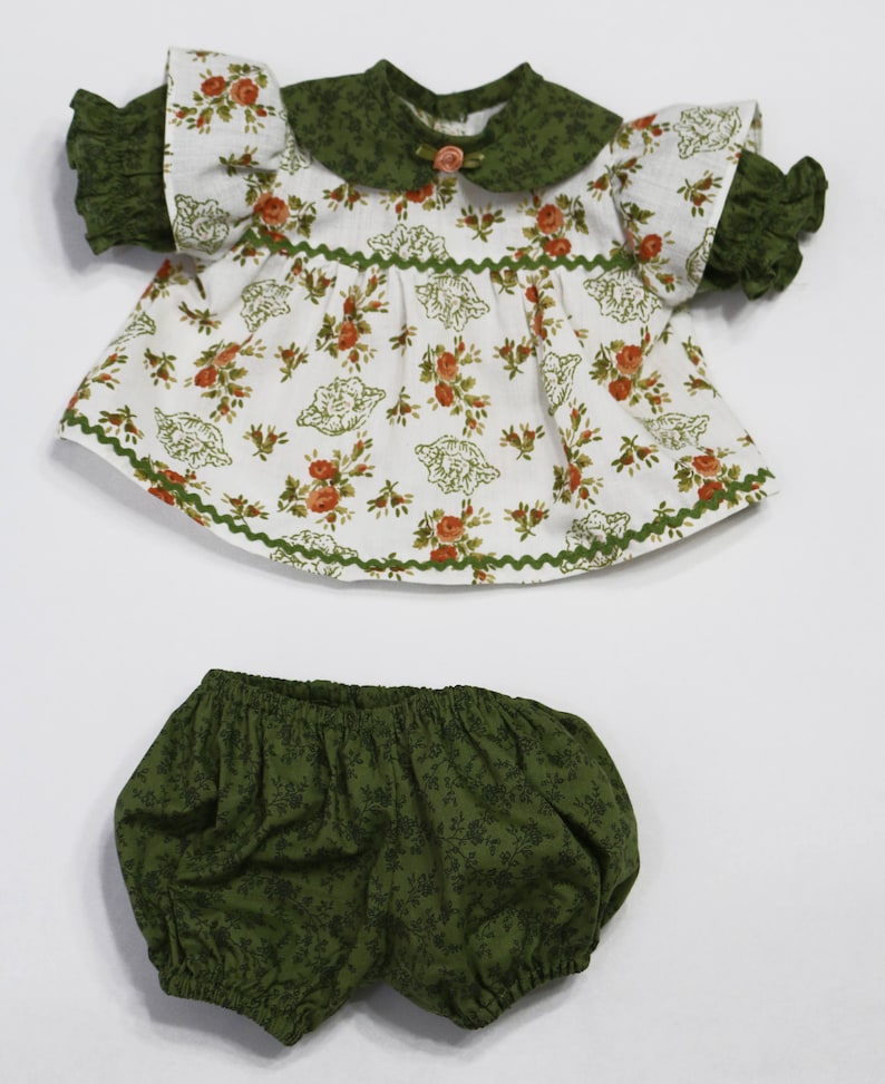 May include: A doll's outfit featuring a white top with a floral pattern in orange and green, a green collar, and matching green ruffled sleeves. The outfit includes green bloomers with a floral design. The outfit is designed for a doll.