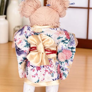 Op de afbeelding: Pop in een bloemenkimono met een grote strik en rode accenten. De pop heeft garen haar gestyled in twee knotjes met decoratieve haarstokjes. De kimono heeft een witte achtergrond met roze en paarse bloempatronen.