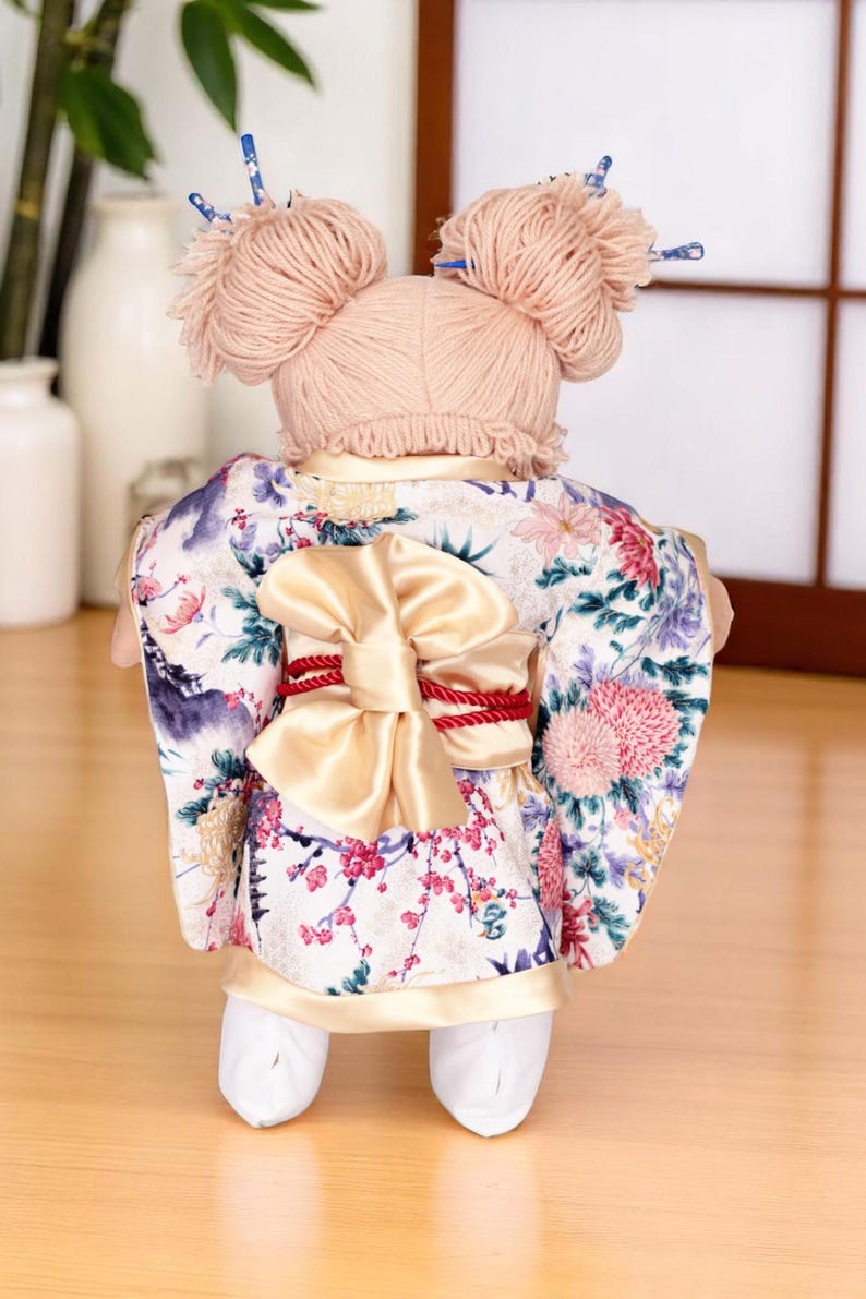 Op de afbeelding: Pop in een bloemenkimono met een grote strik en rode accenten. De pop heeft garen haar gestyled in twee knotjes met decoratieve haarstokjes. De kimono heeft een witte achtergrond met roze en paarse bloempatronen.