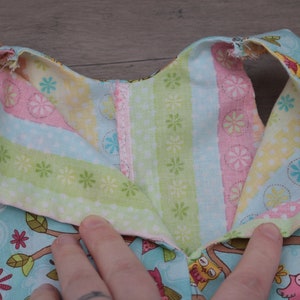 Pode incluir: Close-up da gola de um vestido de beb&eacute;. A gola &eacute; feita de tecido azul claro com um padr&atilde;o de coruja e galho de &aacute;rvore. O interior da gola apresenta tecido listrado em rosa, verde e amarelo. Uma fivela de velcro branca &eacute; vis&iacute;vel.