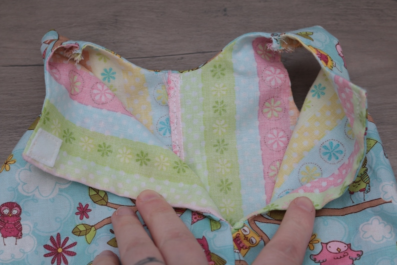 Pode incluir: Close-up da gola de um vestido de beb&eacute;. A gola &eacute; feita de tecido azul claro com um padr&atilde;o de coruja e galho de &aacute;rvore. O interior da gola apresenta tecido listrado em rosa, verde e amarelo. Uma fivela de velcro branca &eacute; vis&iacute;vel.
