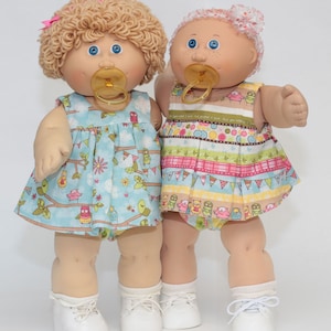 Pode incluir: Duas bonecas de beb&ecirc; em estilo vintage com olhos azuis e chupetas. A boneca da esquerda tem cabelo loiro encaracolado e um vestido azul com estampa de coruja. A boneca da direita tem uma faixa de cabelo rosa e um vestido colorido com um padr&atilde;o listrado. Ambas as bonecas usam sapatos brancos.