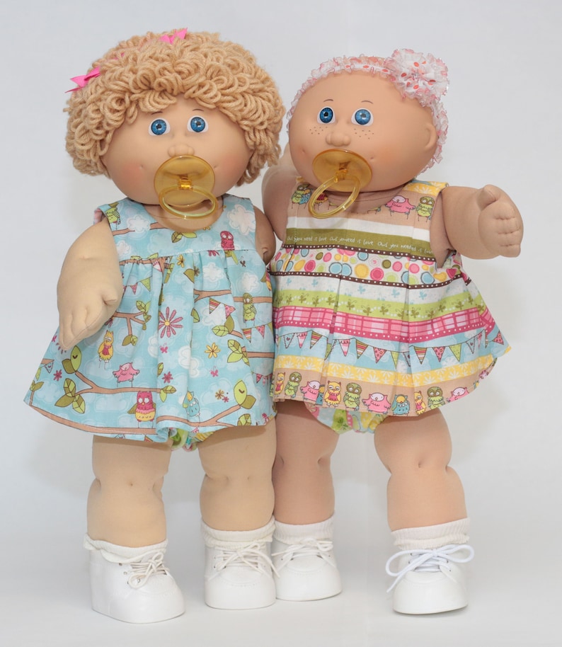 Pode incluir: Duas bonecas de beb&ecirc; em estilo vintage com olhos azuis e chupetas. A boneca da esquerda tem cabelo loiro encaracolado e um vestido azul com estampa de coruja. A boneca da direita tem uma faixa de cabelo rosa e um vestido colorido com um padr&atilde;o listrado. Ambas as bonecas usam sapatos brancos.