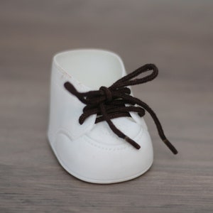 May include: A white baby shoe with brown laces. The shoe has a rounded toe and a simple design.
