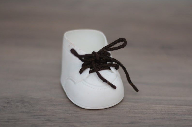 May include: A white baby shoe with brown laces. The shoe has a rounded toe and a simple design.