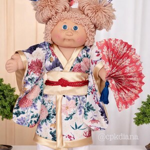 Op de afbeelding: Cabbage Patch Kid pop met een bloemenkimono, rode obi en een rode waaier. De pop heeft lichtbruin garen haar in twee knotjes met haarstokjes. De kimono heeft roze, paarse en witte bloemen. Een verzamelobject.