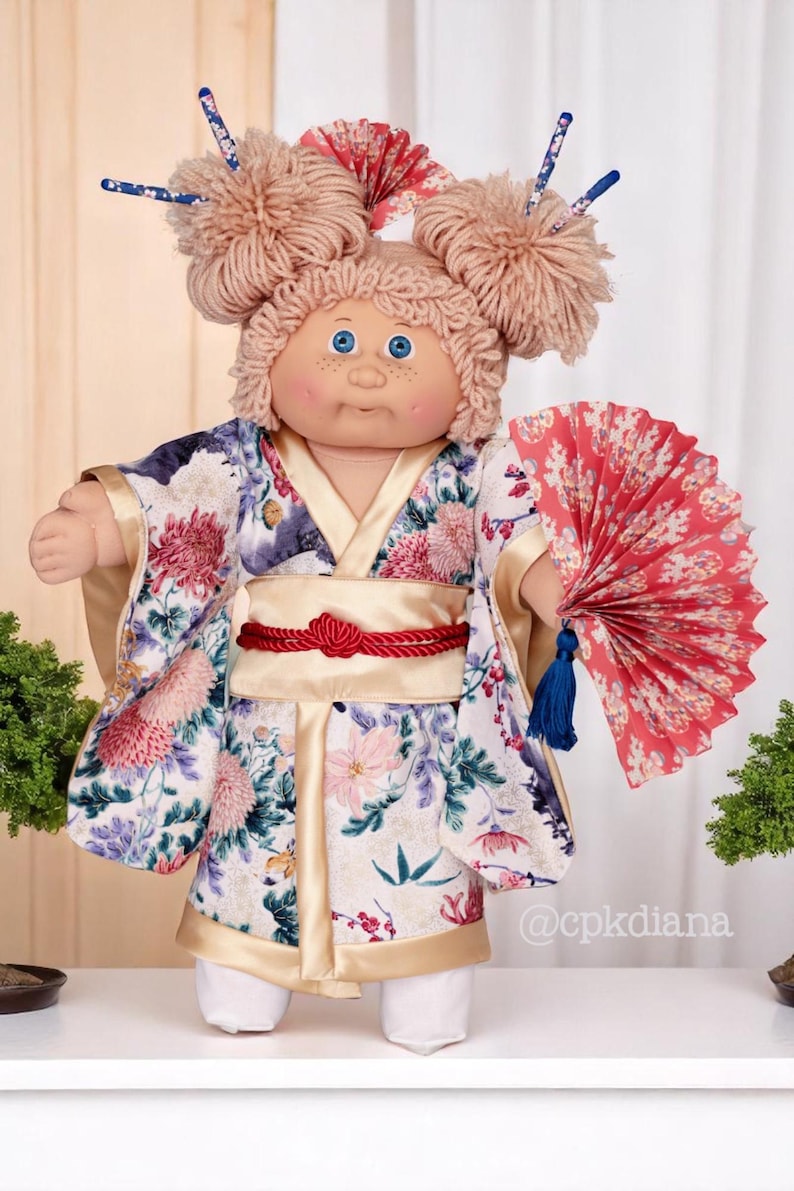 Op de afbeelding: Cabbage Patch Kid pop met een bloemenkimono, rode obi en een rode waaier. De pop heeft lichtbruin garen haar in twee knotjes met haarstokjes. De kimono heeft roze, paarse en witte bloemen. Een verzamelobject.