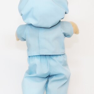 K&ouml;nnte beinhalten: Eine hellblaue Puppe in einem OP-Kittel-Outfit, einschlie&szlig;lich einer M&uuml;tze, einem kurz&auml;rmeligen Hemd und einer Hose. Die Arme der Puppe sind sichtbar, und die Hose hat einen R&uuml;schensaum.