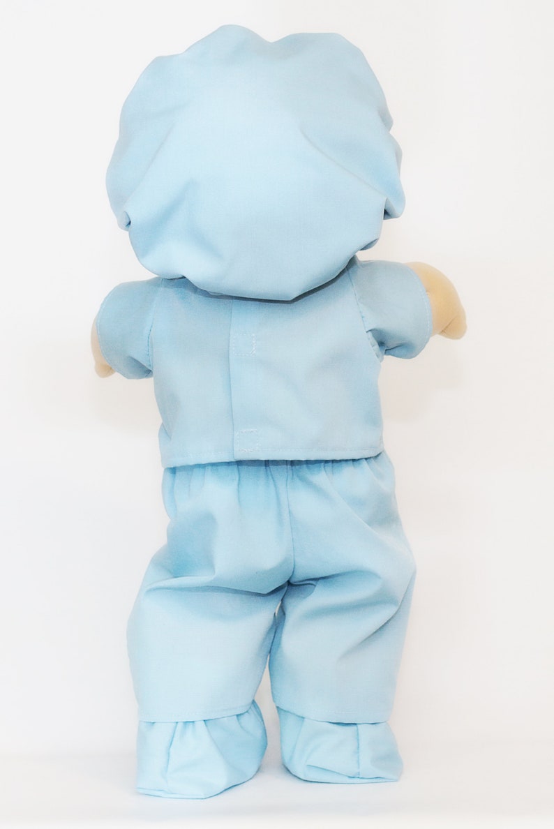 K&ouml;nnte beinhalten: Eine hellblaue Puppe in einem OP-Kittel-Outfit, einschlie&szlig;lich einer M&uuml;tze, einem kurz&auml;rmeligen Hemd und einer Hose. Die Arme der Puppe sind sichtbar, und die Hose hat einen R&uuml;schensaum.