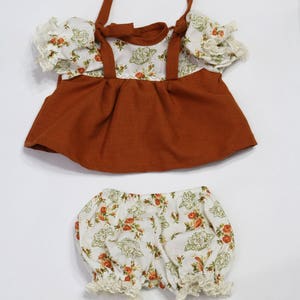 May include: A two-piece doll outfit featuring a rust-colored top with floral print accents and matching bloomers. The top has tie straps and ruffled sleeves. The bloomers are white with a floral pattern and lace trim.