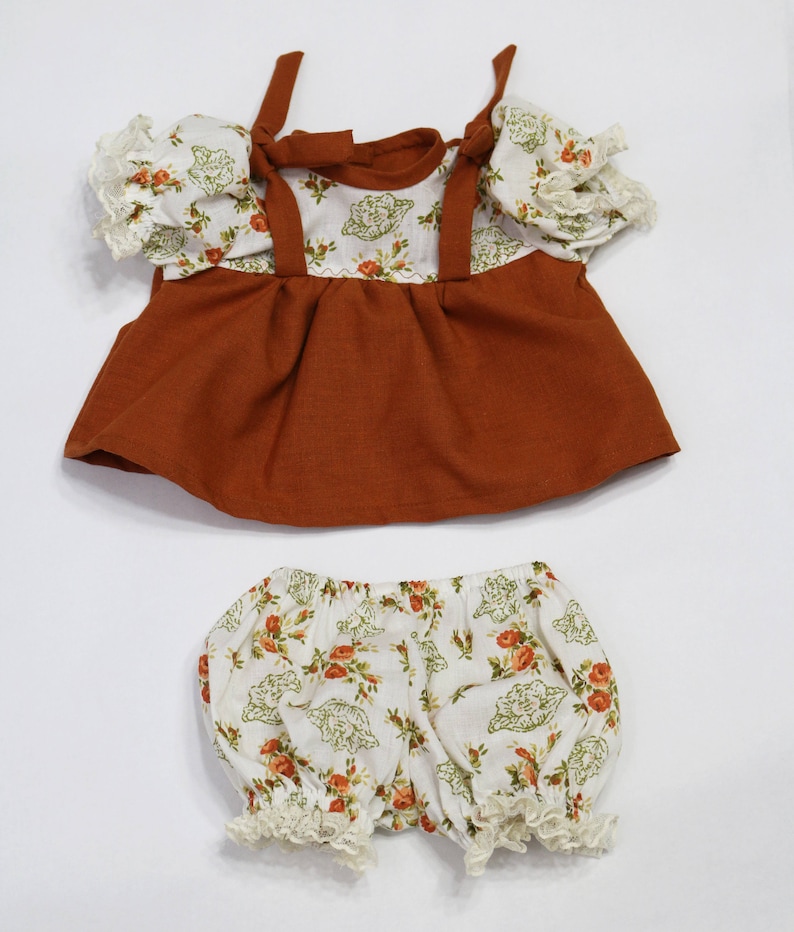 May include: A two-piece doll outfit featuring a rust-colored top with floral print accents and matching bloomers. The top has tie straps and ruffled sleeves. The bloomers are white with a floral pattern and lace trim.