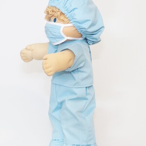 K&ouml;nnte beinhalten: Eine Cabbage Patch Kid-Puppe in einem hellblauen OP-Anzug, einschlie&szlig;lich M&uuml;tze, Maske, Oberteil und Hose. Die Puppe hat blonde Locken und blaue Augen. Das Outfit hat wei&szlig;e Verzierungen und ist aus einem weichen Stoff gefertigt.