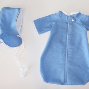 May include: A baby blue sleep sack and matching bonnet set. The sleep sack has short sleeves, a full-length zip, and white trim. The bonnet has a ruffled brim and ties with white cords. The set is made of soft fabric.