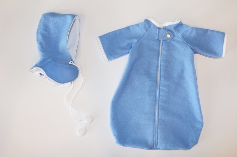May include: A baby blue sleep sack and matching bonnet set. The sleep sack has short sleeves, a full-length zip, and white trim. The bonnet has a ruffled brim and ties with white cords. The set is made of soft fabric.