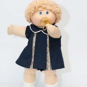 May include: A vintage doll with blonde, curly hair, blue eyes, and a pacifier. The doll wears a navy blue dress with a white collar and floral accents, white socks, and white shoes. The doll is standing against a white background.