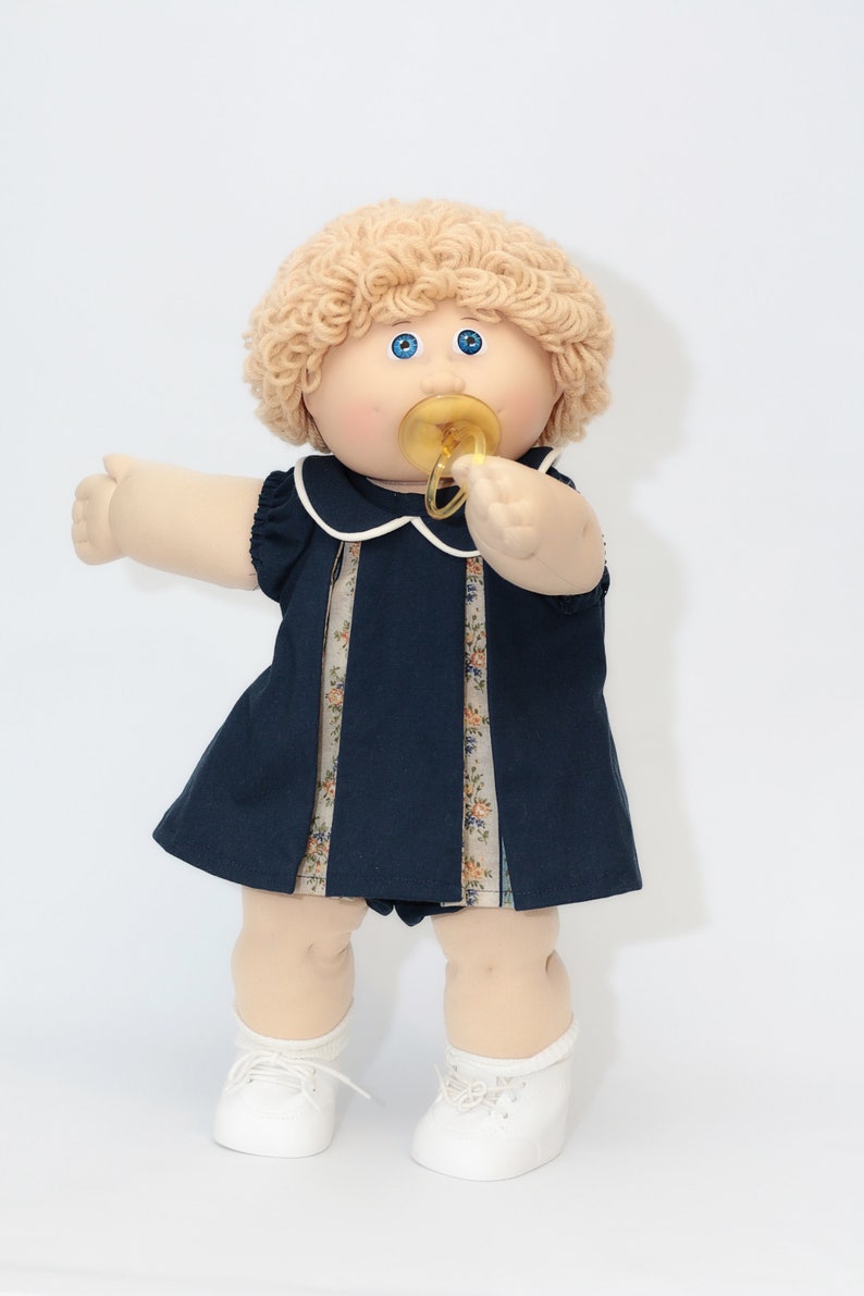 May include: A vintage doll with blonde, curly hair, blue eyes, and a pacifier. The doll wears a navy blue dress with a white collar and floral accents, white socks, and white shoes. The doll is standing against a white background.